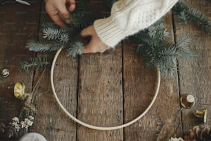Hands in cozy sweater making modern christmas wreath with fir branches, herbs, bells, round wooden hoop on rustic table. Atmospheric moody image. Making xmas boho wreath