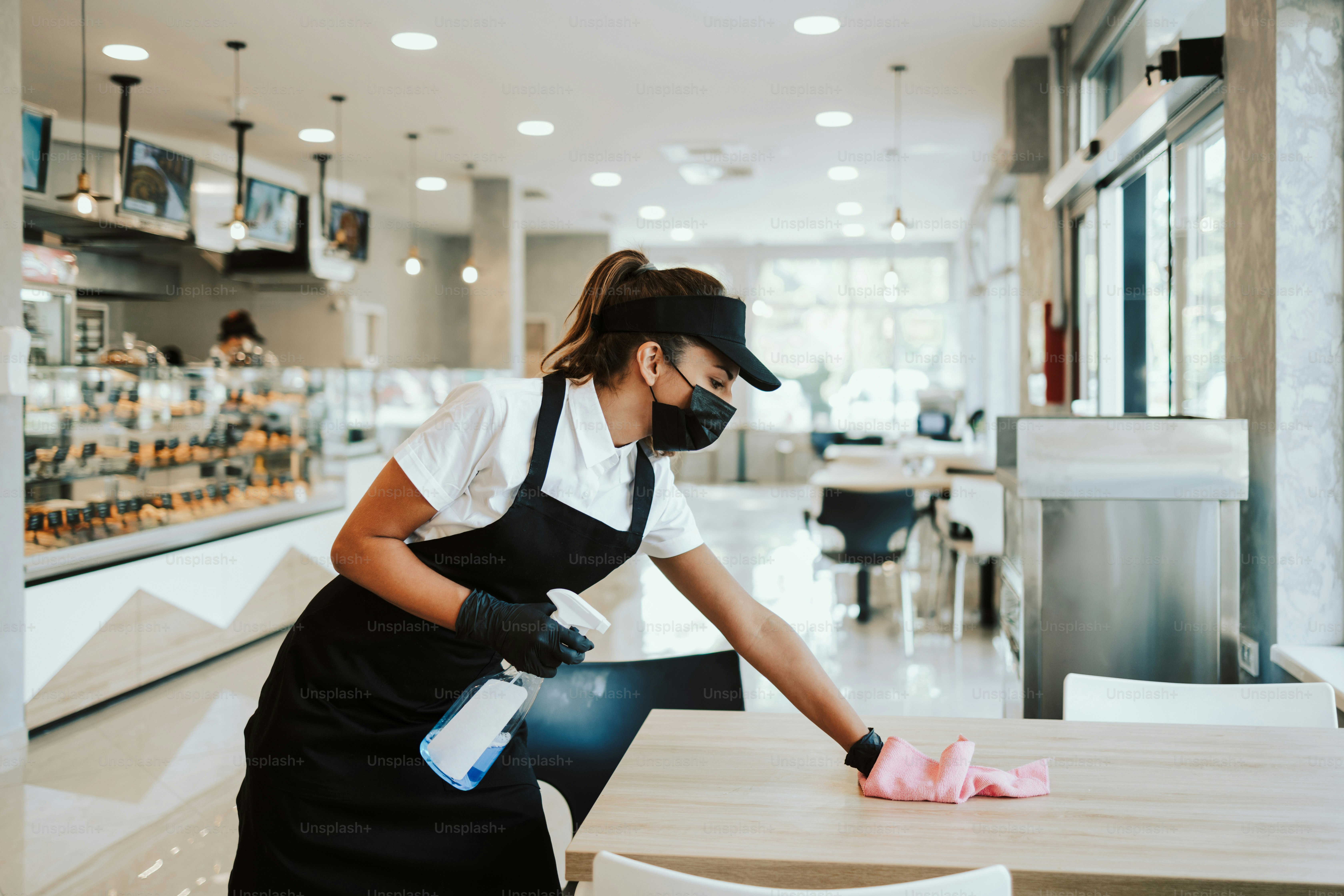 Beautiful woman working bakery or fast food restaurant. She is cleaning and disinfecting tables against Coronavirus pandemic disease. She is wearing protective face masks, gloves and face shield.