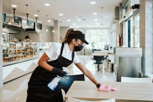 Schöne Frau, die in einer Bäckerei oder einem Fast-Food-Restaurant arbeitet. Sie reinigt und desinfiziert Tische gegen die Coronavirus-Pandemie. Sie trägt Schutzmasken, Handschuhe und Gesichtsschutz.