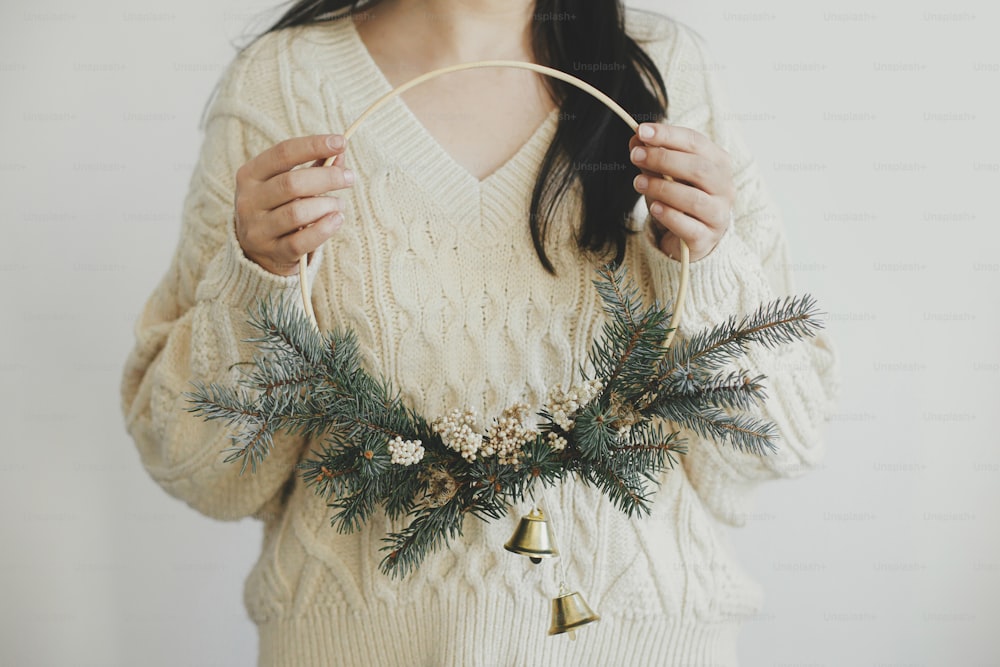 Mulher elegante no suéter aconchegante que segura a coroa de Natal moderna no fundo branco da parede. Feliz Natal e Boas Festas! Coroa minimalista de xmas boho com sinos nas mãos. Tempo atmosférico