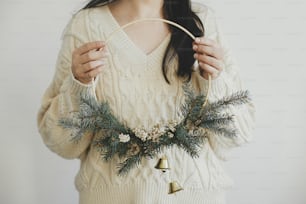 하얀 벽 배경에 현대 크리스마스 화환을 들고 아늑한 스웨터를 입은 세련된 여성. 메리 크리스마스와 해피 홀리데이! 손에 종소리가 있는 미니멀리스트 크리스마스 보호 화환. 대기 시간