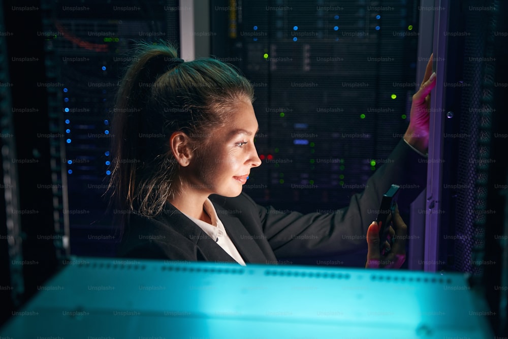 Photo of young smiling woman checking data on cellphone while working with supercomputer in research center