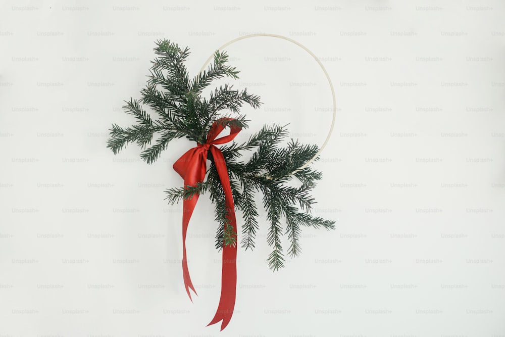 白い壁の背景にぶら下がっているモダンなミニマリストのクリスマスリース。メリークリスマスとハッピーホリデー!モミの枝と赤い弓とスタイリッシュなクリスマスリース、隔離