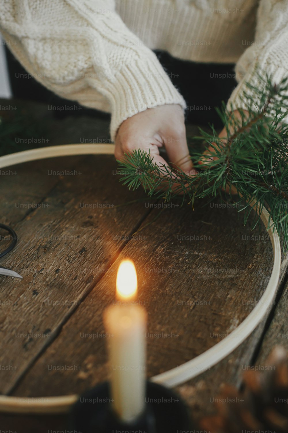 Hands making modern christmas wreath on rustic table with fir branches, ribbons, round wooden hoop, scissors, thread, candle. Atmospheric moody image. Winter holidays preparation