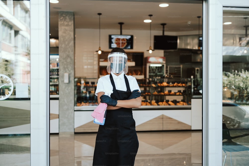 Beautiful woman with face shield and protective mask working bakery or fast food restaurant. She is standing in front of shop and holding disinfectants with arms crossed. Covid-19 safety measures.