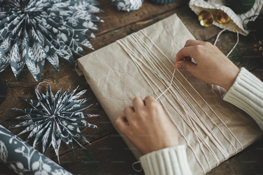 Envolver el regalo de Navidad. Manos envolviendo un elegante regalo de Navidad simple en papel artesanal con cuerda sobre una mesa de madera rústica con estrellas de papel azul. Imagen atmosférica y temperamental, estilo nórdico. ¡Feliz Navidad!