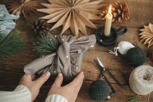 Hände mit Weihnachtsgeschenk verpackt in modernen festlichen Stoff auf rustikalem Holztisch mit Ornamenten. Atmosphärisches stimmungsvolles Bild, nordischer Stil. Frohe Weihnachten! Furoshiki Wrap, Zero Waste Urlaub