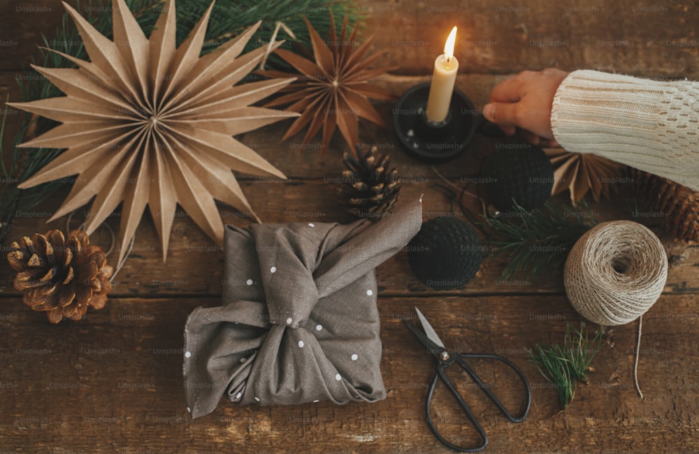 Christmas advent. Hand holding candle and stylish christmas gift wrapped in fabric, scissors, paper star, ornaments on rustic wooden table. Atmospheric moody image, nordic style. Merry Christmas!