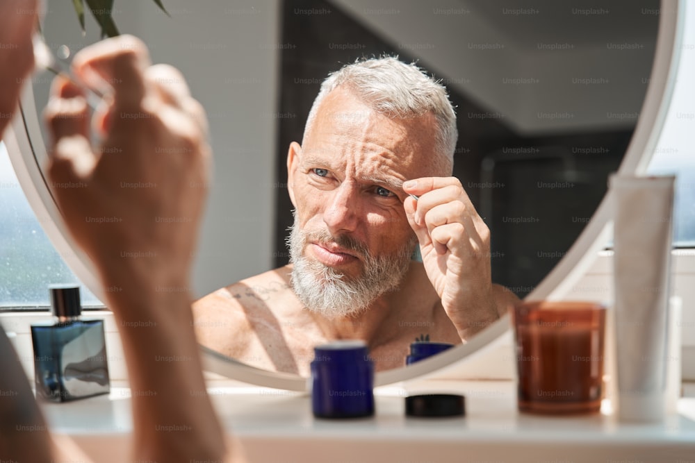 Mature man tweezing eyebrows on his face while looking at mirror in bathroom. Concept of face skin care and hygiene. Domestic lifestyle. Grey haired european male pensioner with tattoos