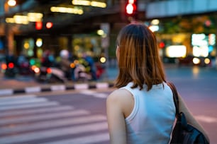 Portrait of Young beautiful Asian woman walking on street crosswalk in the city and looking at crowd of people and illuminated night lights. Pretty girl enjoy urban outdoor lifestyle and city night life.