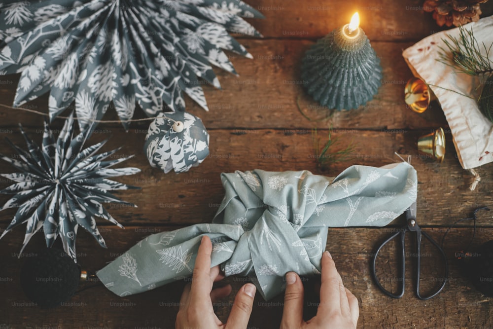 Hands holding christmas gift wrapped in fabric on rustic wooden table with candle, ornaments, blue paper stars. Atmospheric moody image, nordic style. Merry Christmas! Furoshiki wrap flat lay