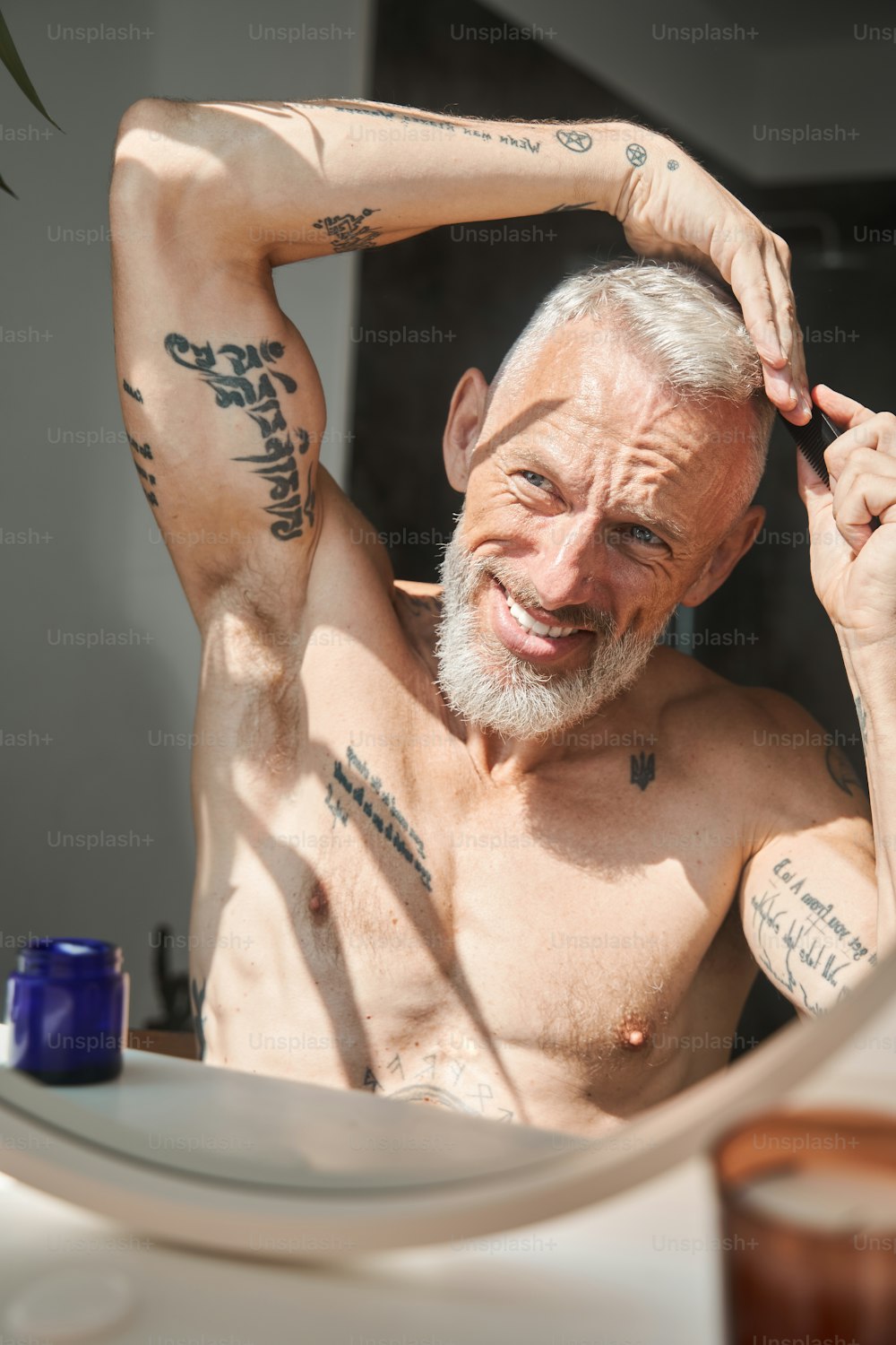 Mature man combing hair on his head while looking at mirror in bathroom. Concept of skin care and hygiene. Domestic lifestyle. Smiling grey haired european male pensioner with tattoos