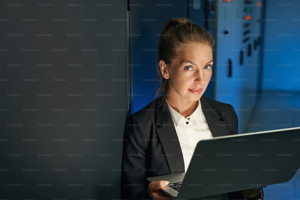 サーバールームでスーパーコンピュータを操作しながらノートパソコンを持ち、カメラを見つめる女性データエンジニアの写真