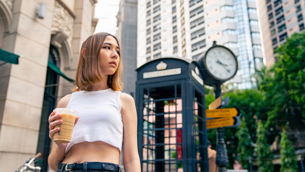 Portrait of Young beautiful Asian woman in casual clothing sitting on outdoor bench in the city and drinking iced coffee with looking crowd of people walking street. Pretty girl enjoy urban lifestyle and city life.