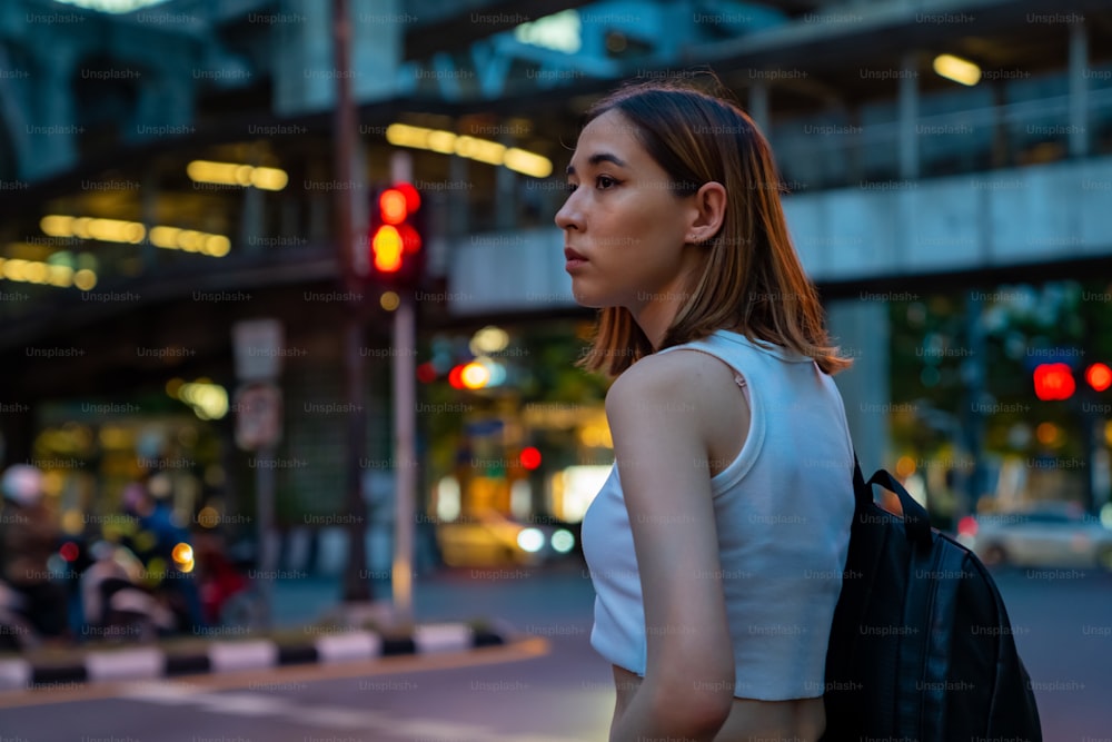 Ritratto di giovane bella donna asiatica che cammina sulle strisce pedonali della città e guarda la folla di persone e le luci notturne illuminate. La bella ragazza ama lo stile di vita urbano all'aperto e la vita notturna della città.