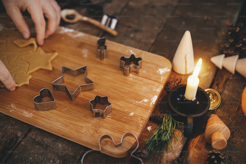 Preparazione dei tradizionali biscotti di pan di zenzero natalizi su un tavolo rustico con candela, spezie, decorazioni. Immagine lunatica. Tagliare a mano l'impasto di pan di zenzero con taglierine metalliche natalizie su tavola di legno