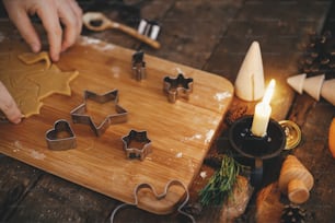 촛불, 향신료, 장식으로 소박한 테이블에 전통적인 크리스마스 진저 브레드 쿠키를 만듭니다. 무디 이미지. 나무 보드에 크리스마스 금속 절단기로 진저 빵 반죽을 손으로 자르십시오.