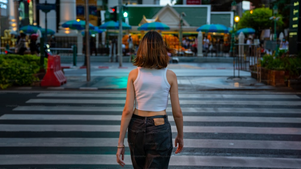 街の横断歩道を歩いていて、人々の群衆と照らされた常夜灯を見ている若い美しいアジア人女性の肖像画。かわいい女の子は、都会のアウトドアライフスタイルと街のナイトライフを楽しんでいます。