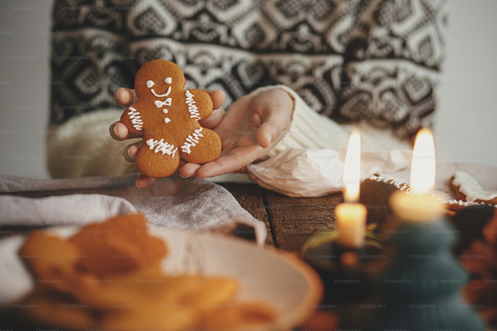 Hands holding decorated gingerbread man christmas cookie on background of rustic table with napkin, candle, decorations. Moody image. Woman making stylish christmas gingerbread cookies