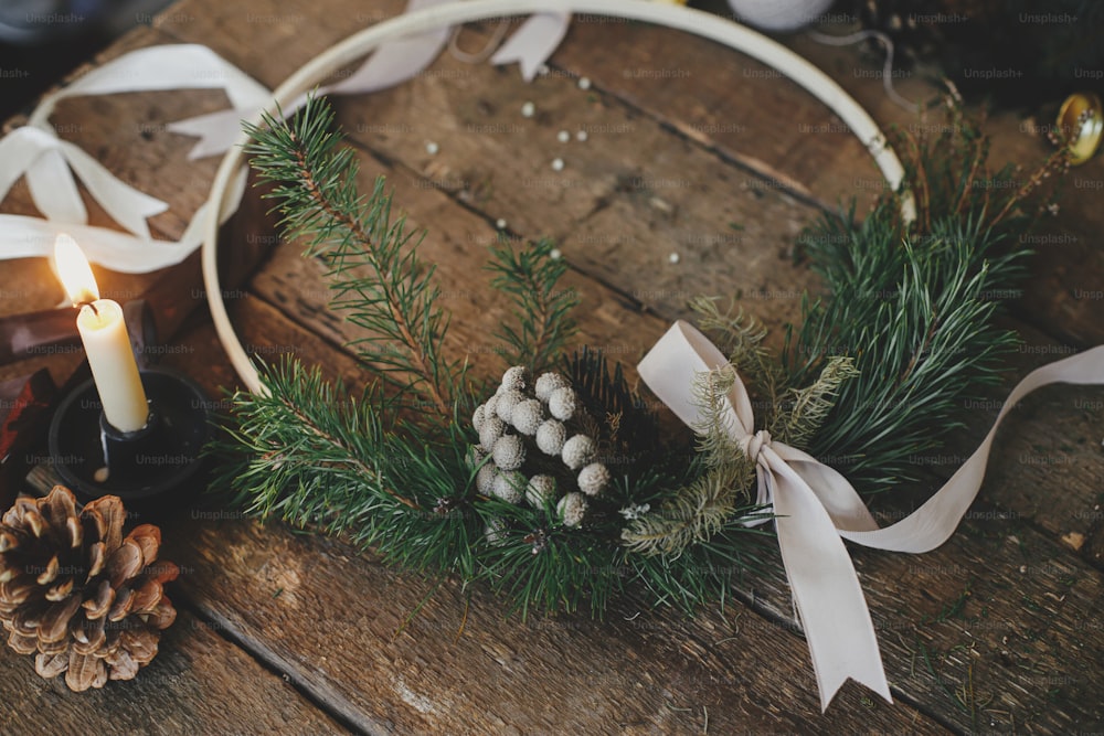 素朴な木製のテーブルにスタイリッシュでモダンなクリスマスリース、リボン、キャンドル、糸、松ぼっくり。メリークリスマスとハッピーホリデー!冬休みの準備、雰囲気イメージ