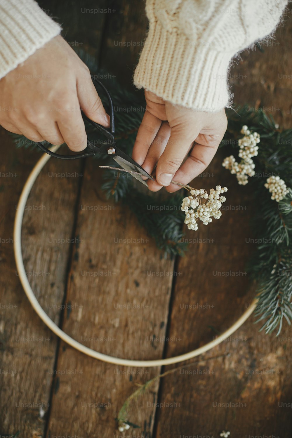 Hands in cozy sweater cutting herb with scissors for modern boho wreath with fir branches and wooden hoop on rustic table. Atmospheric moody image. Making stylish christmas wreath