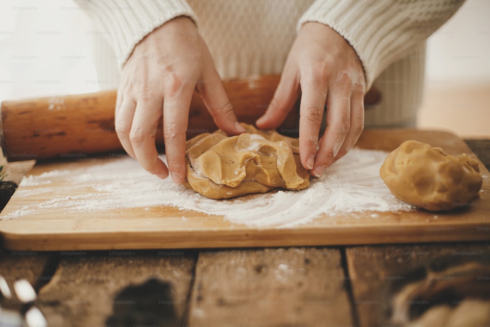 麺棒と小麦粉で木の板の上でクリスマスの伝統的なクッキーのためにジンジャーブレッド生地をこねる手。素朴なテーブルでクリスマスジンジャーブレッドクッキーを作る