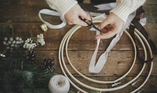 Making stylish minimalist christmas wreath. Hands cutting ribbon with scissors for modern boho wreath with fir branches, brunia herb, wooden hoop on rustic table. Atmospheric moody image