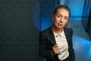 黒いスーツを着た自信に満ちた女性システム管理者が、データセンターの部屋のカメラを背景に見ている