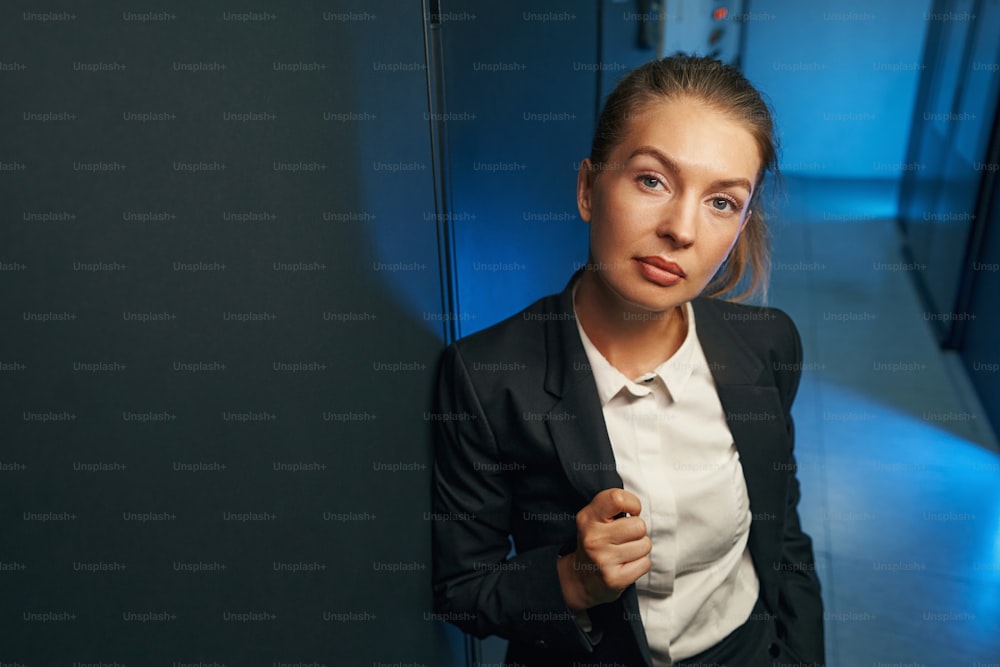 Confident female system administrator in black suit looking at camera in room of datacenter on background
