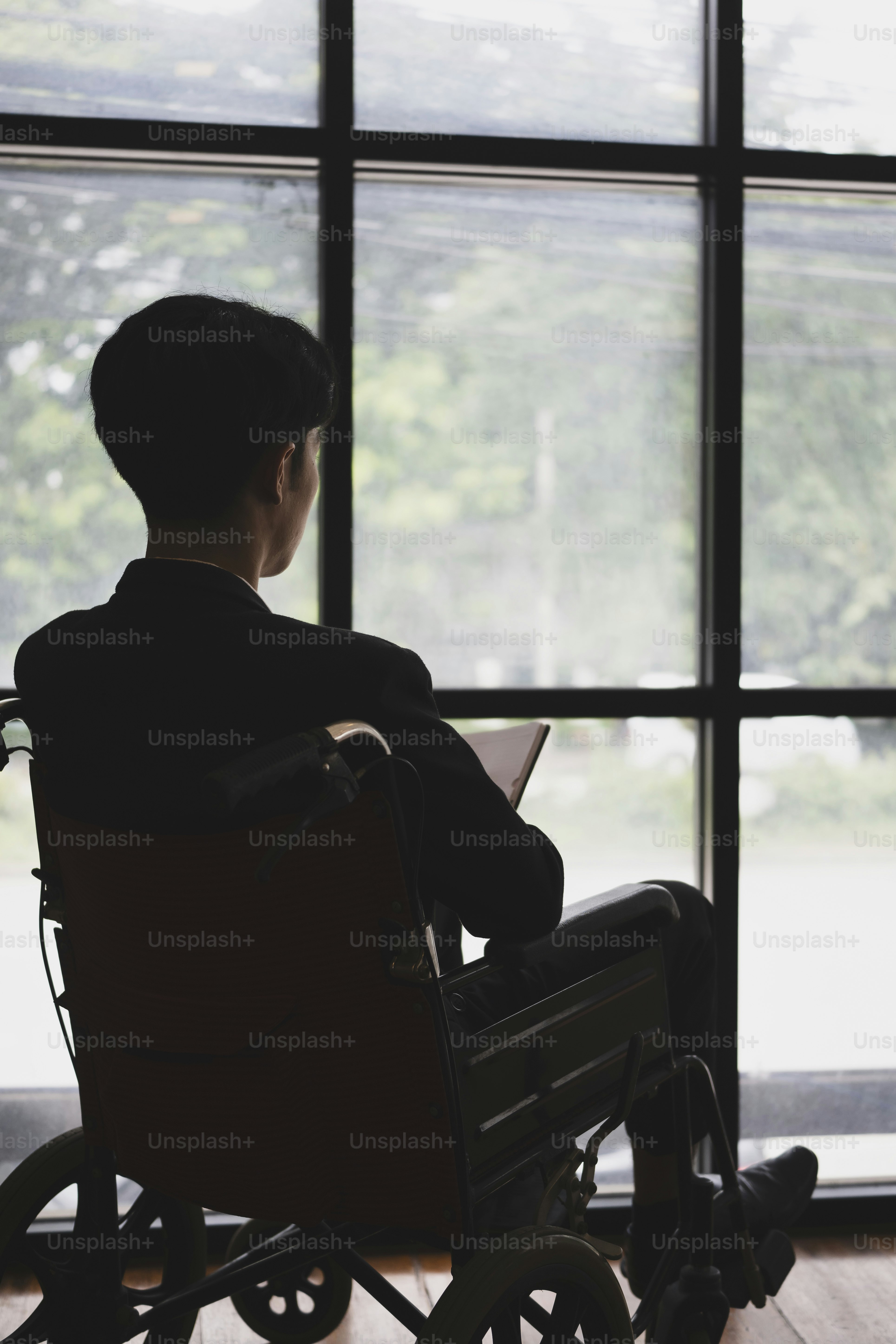 Rear view of thoughtful businessman in wheelchair sitting in office room and looking out of window.