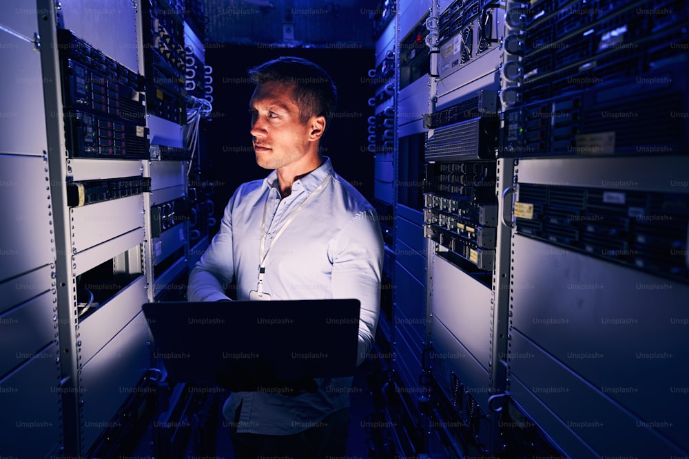 Serious IT technician with laptop monitoring network servers in colocation data center
