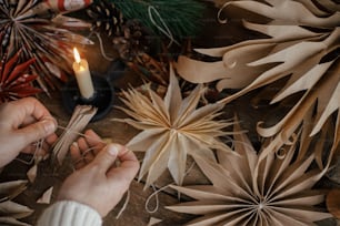 Making stylish Christmas stars. Hands holding folded craft paper on background of handmade sweden stars, thread, candle, scissors on rustic wood. Process of making festive decor.