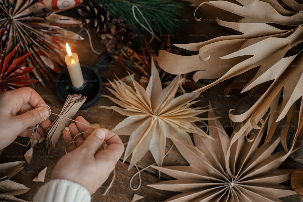 Realizzare eleganti stelle di Natale. Mani che tengono carta artigianale piegata su sfondo di stelle svedesi fatte a mano, filo, candela, forbici su legno rustico. Processo di realizzazione di decorazioni festive.