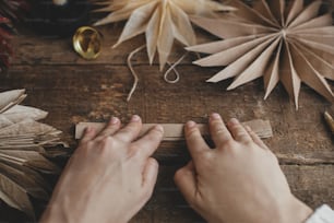 Making stylish Christmas stars. Hands folding craft paper on background of handmade sweden stars, thread, candle, scissors on rustic wood. Process of making festive decor. Holiday preparations