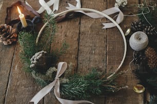 Modern christmas wreath on rustic wooden background. Stylish boho wreath with brunia on rustic table with ribbons, candle, thread, pine cones. Atmospheric image. Merry Christmas and Happy holidays!