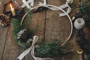 Corona navideña moderna sobre fondo rústico de madera. Elegante corona boho con brunia sobre mesa rústica con cintas, vela, hilo, piñas. Imagen atmosférica. ¡Feliz Navidad y Felices Fiestas!