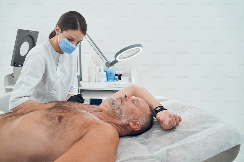 Une femme médecin portant un masque médical marquant l’aisselle du client avant l’intervention tandis qu’un homme est allongé sur un lit de repos avec les yeux fermés