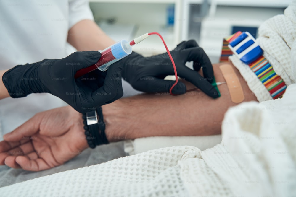 助手が男性の腕に針を挿入している間、血液の入った試験管を保持する黒い手袋をはめた医師の手の接写