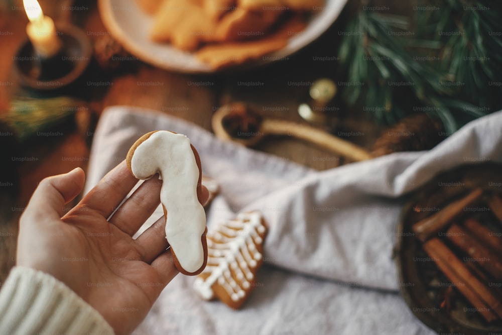 Hand holding decorated gingerbread cookie candy cane on background of rustic table with napkin, candle, decorations. Moody image. Woman making stylish christmas gingerbread cookies