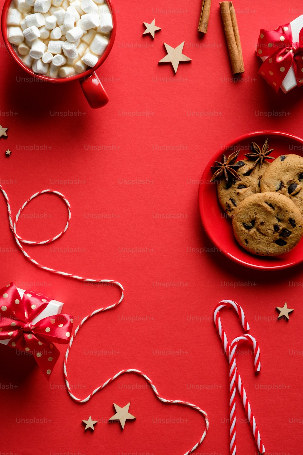 Composition de Noël avec des biscuits à l’avoine, des cannes de bonbon, des bâtons de cannelle, du chocolat chaud aux guimauves, des décorations d’étoiles en bois sur fond rouge. Pose à plat, vue de dessus