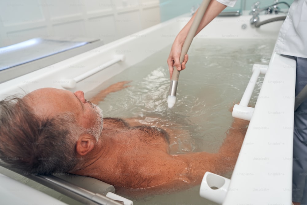 Male person lying in bathtub while cosmetologist performing hydromassage with special device