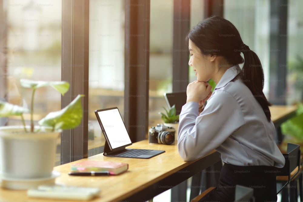 Vista lateral, hermosa joven empresaria asiática que trabaja en la tableta, mirando en la pantalla, pensando en crear contenido en línea, sentada en la cafetería.