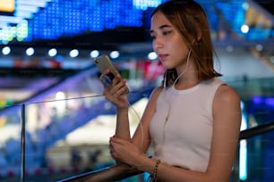 Beautiful Asian woman walking down city street with listening to the music from earphones on smartphone application and looking at illuminated city street night lights. Pretty girl enjoy urban outdoor lifestyle at night