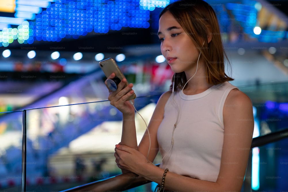 Schöne asiatische Frau, die die Stadtstraße entlang geht, die Musik von Kopfhörern auf Smartphone-Anwendung hört und die beleuchteten Nachtlichter der Stadt betrachtet. Hübsches Mädchen genießt urbanen Outdoor-Lifestyle in der Nacht