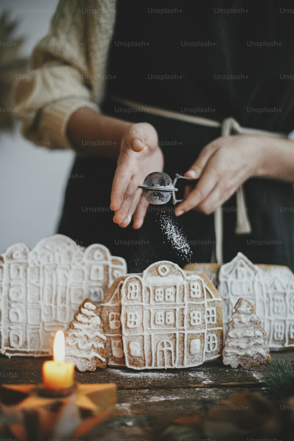 Woman in apron sprinkling sugar powder on christmas gingerbread houses on rustic wooden table. Atmospheric moody image. Christmas holiday preparation and traditions. Decorating cookies