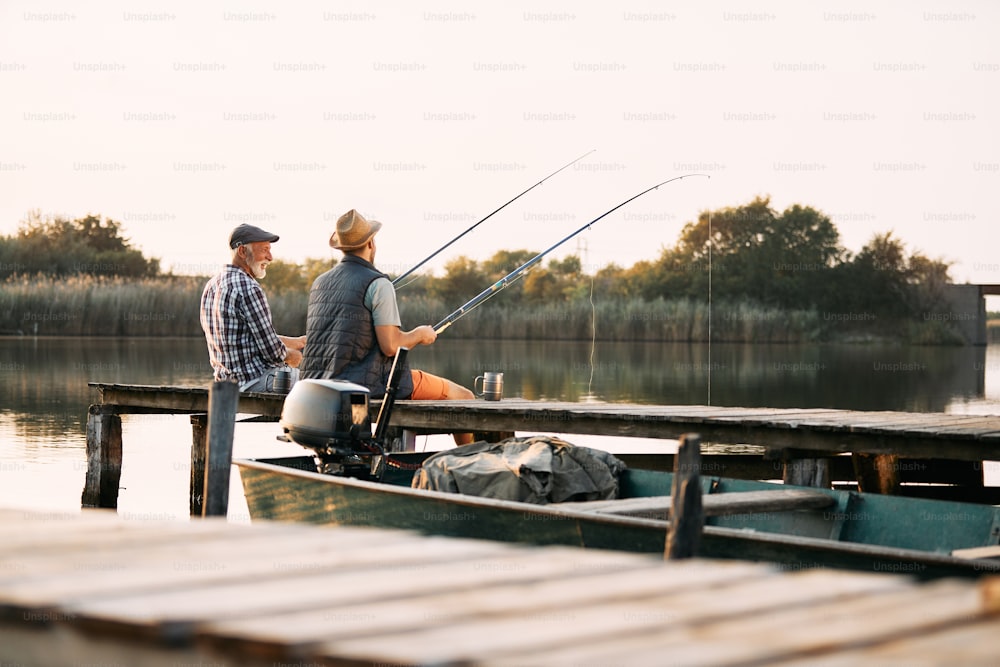 Vista posteriore dell'uomo e di suo padre anziano che pescano da un molo mentre trascorrono del tempo insieme nella natura.
