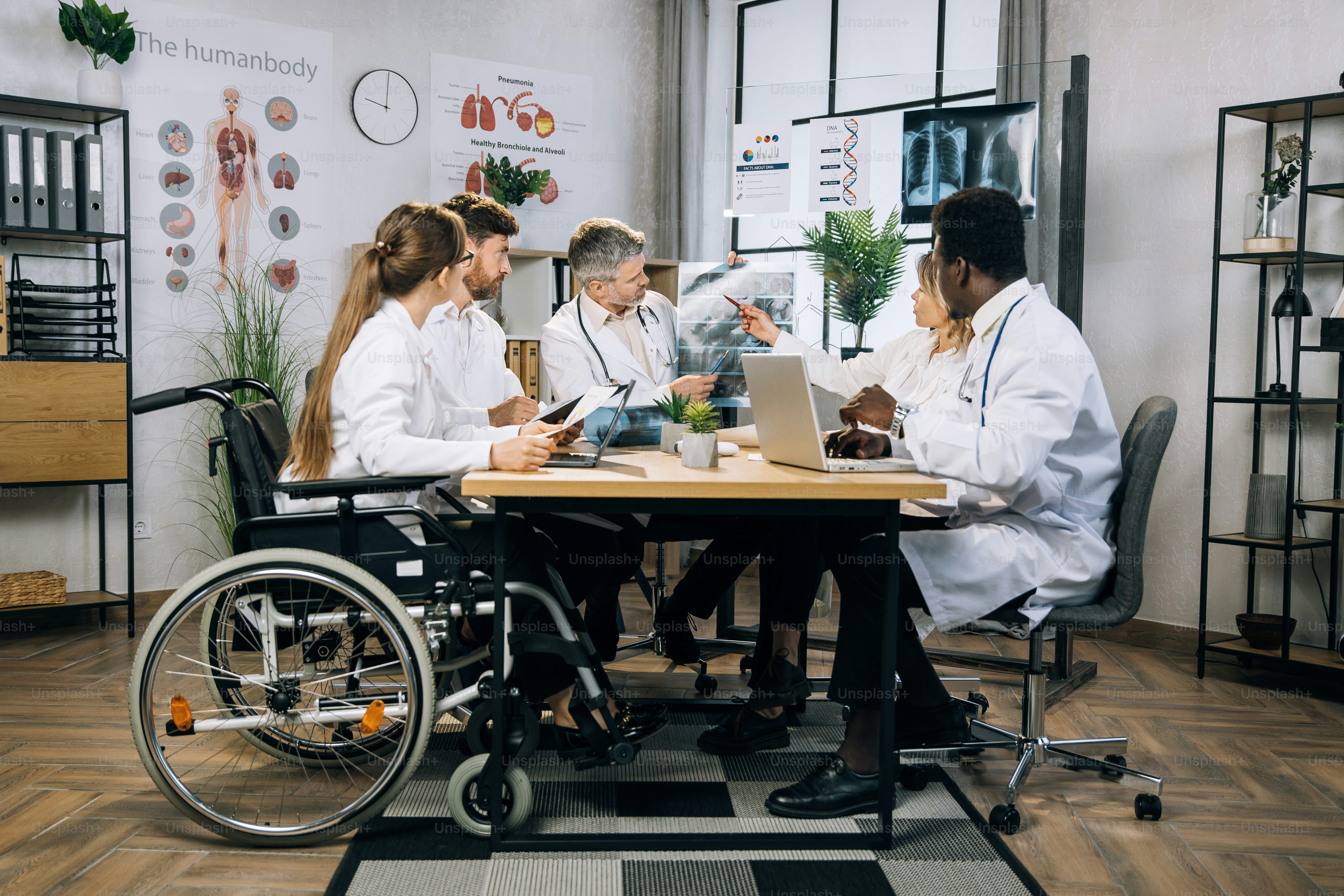 Grupo de cinco trabalhadores médicos multiétnicos discutindo os resultados da varredura de raios-x enquanto estão sentados juntos na sala de escritório. Médicos qualificados e seu colega em cadeira de rodas tendo reunião de trabalho dentro de casa.