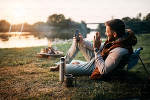 Happy man using mobile phone and waving while talking to his wife and camping in nature. Copy space.