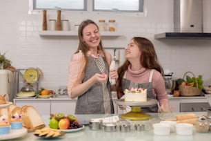 Expectant mother and daughter decorating cupcakes on a kitchen table, having a good time together while eating freshly baked pastries