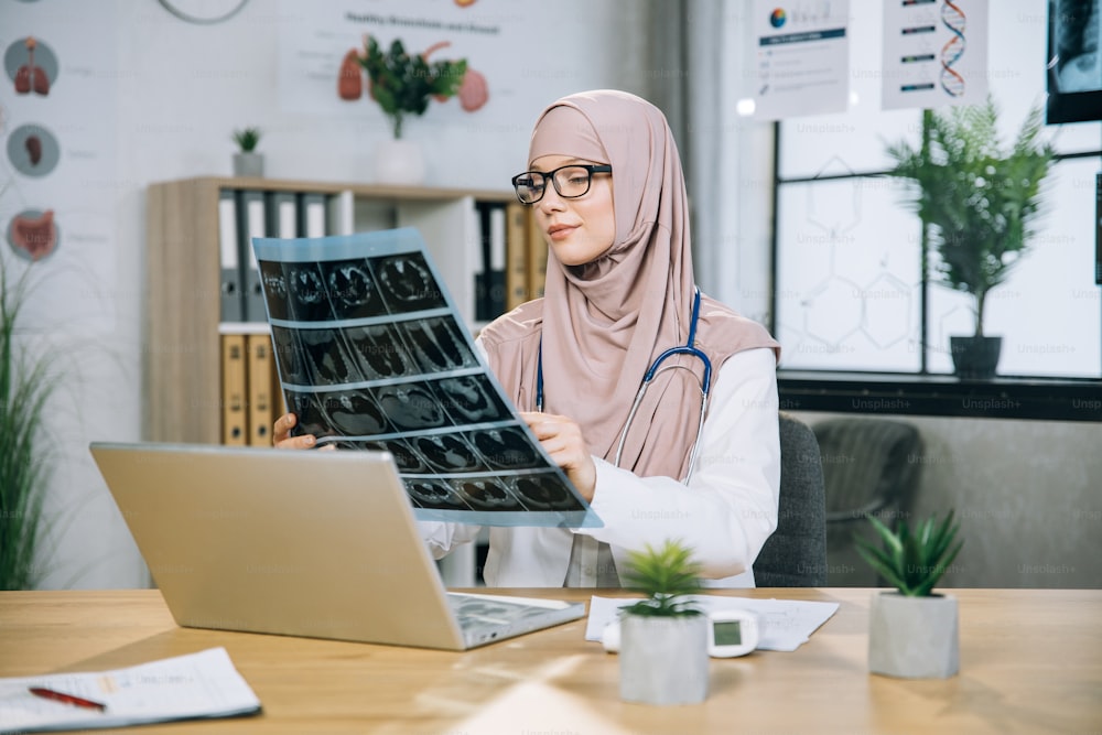 Praticante competente do sexo feminino em hijab examinando os resultados da varredura de raios-x enquanto está sentado à mesa com laptop sem fio. Médico árabe à procura de forma de tratamento do paciente.